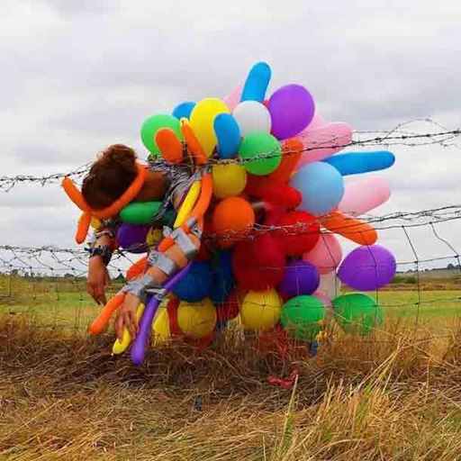A person wearing an outfit made out of colourful balloons flops forward over a barbed wire fence in a field