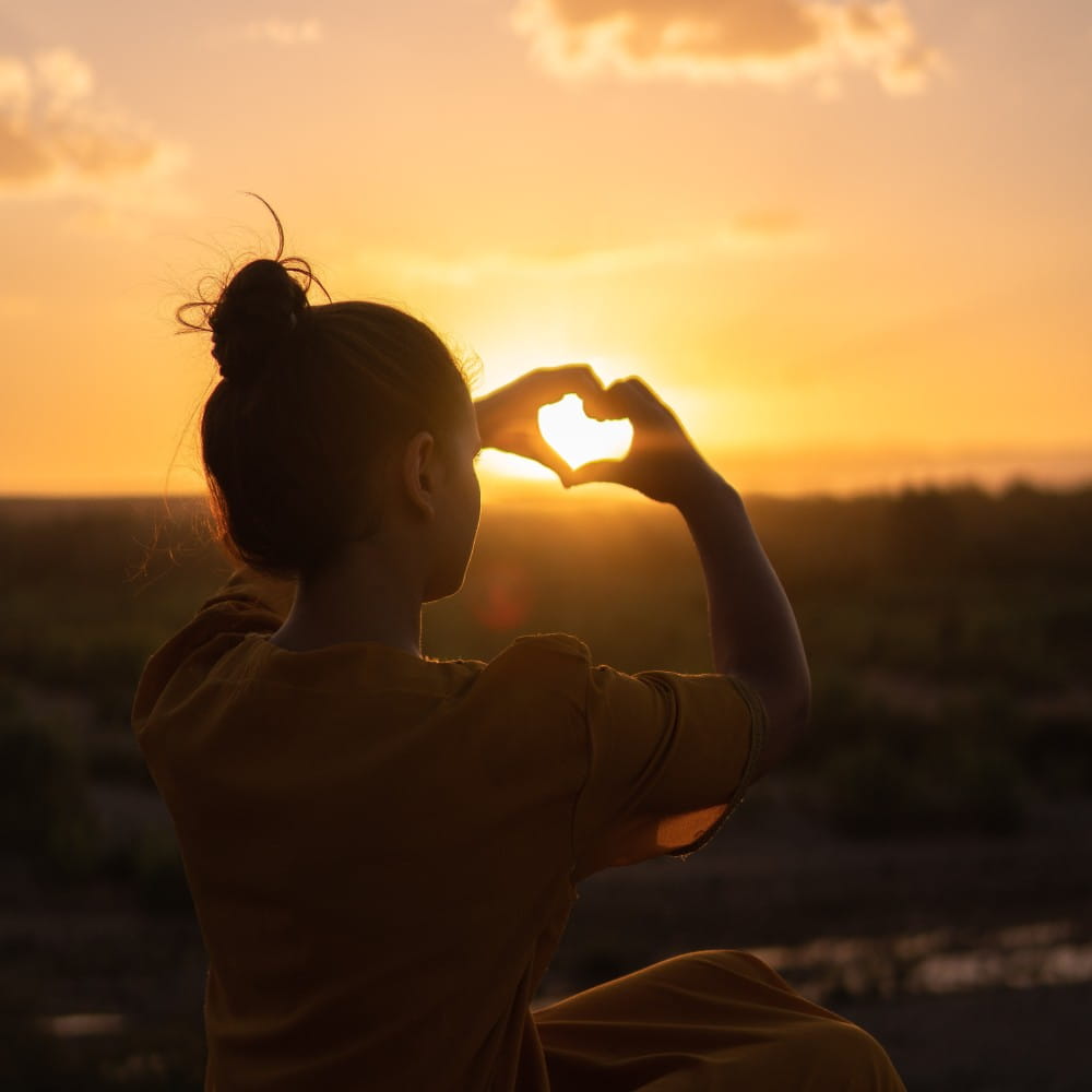 Woman sitting on stone, holding up her arms to make a heart around the sunset in the distance.