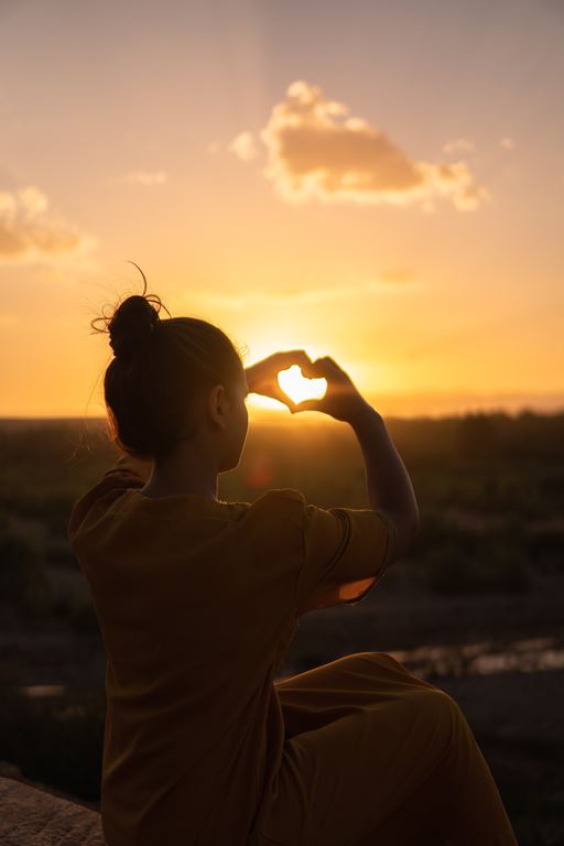 Woman sitting on stone, holding up her arms to make a heart around the sunset in the distance