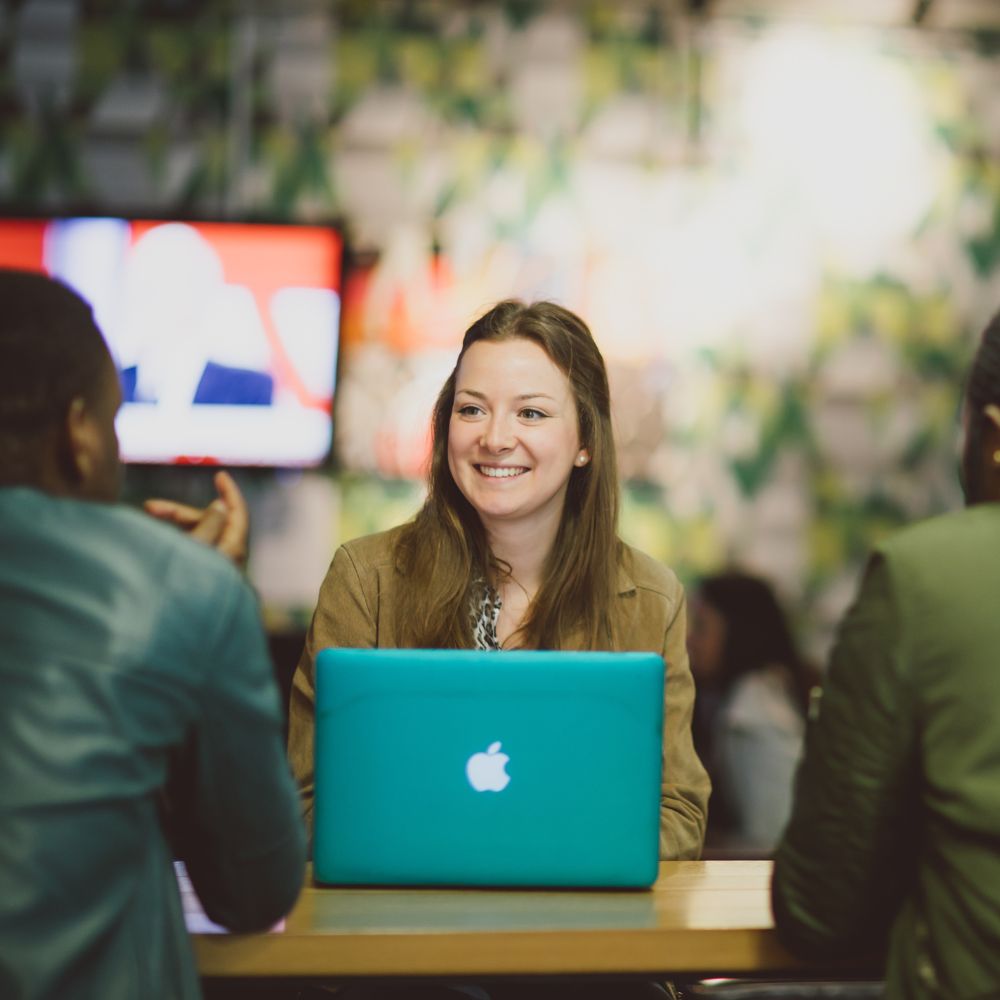 A young woman smiles, as she listens to a colleague over the top of her laptop