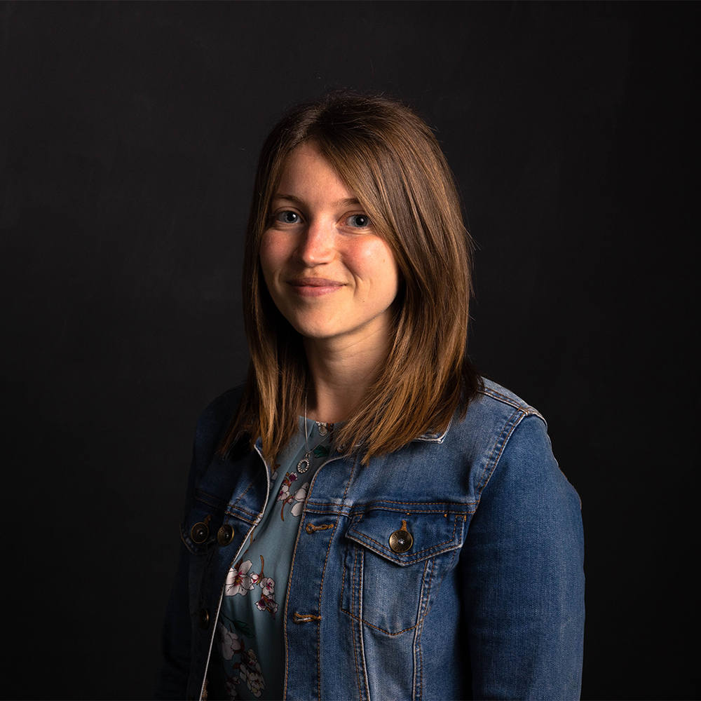 Hannah Crago, Open Research Development Librarian, stands looking towards the camera smiling with her shoulder length hair down.