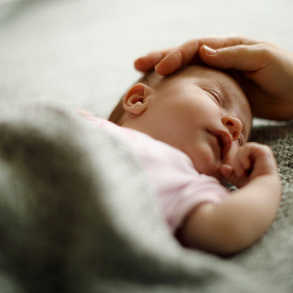A small baby sleeping under a blanket, with an adult's hand stroking their head.