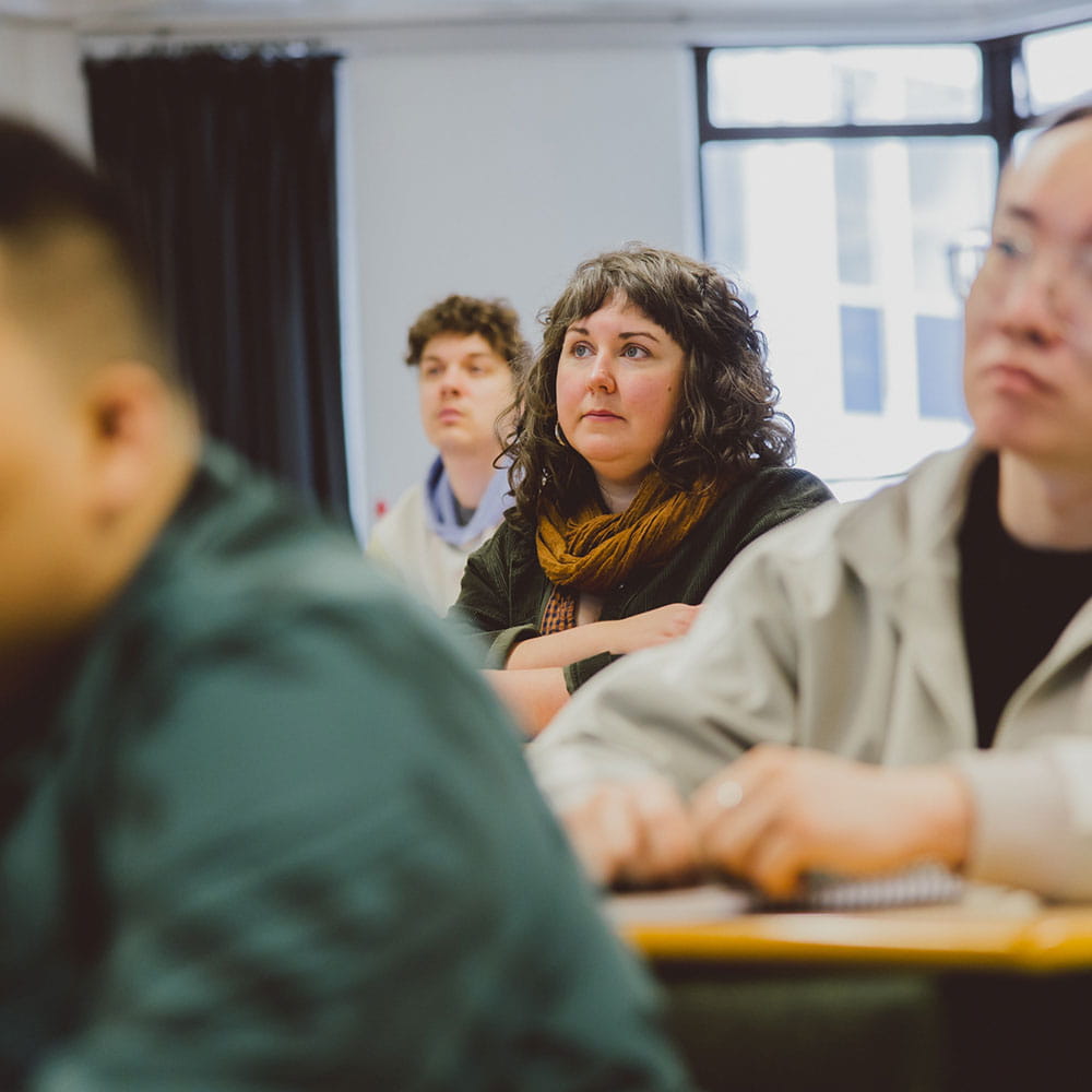 Group of students in seminar room. The students in the foreground are blurred. the student in the background is in focus and looks interested in the discussion in the room.