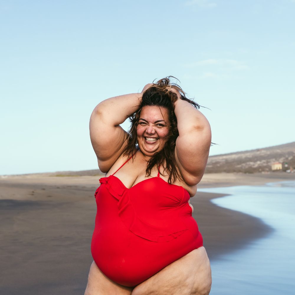 A young plus size woman, wearing a vibrant red swim suit, smiles on a beach
