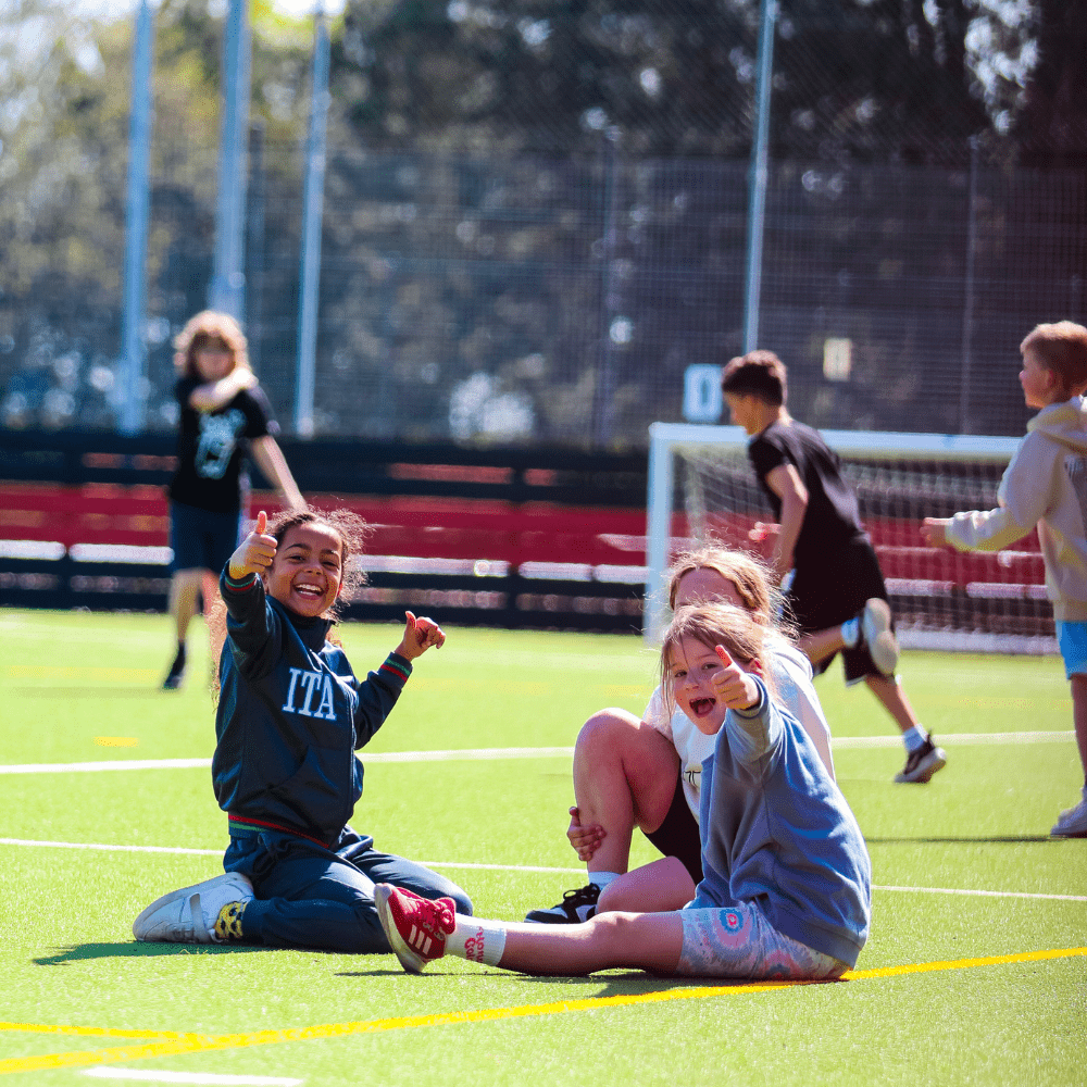 Outdoors -  three children sitting on the floor smilling, in backgound three other children playing football 