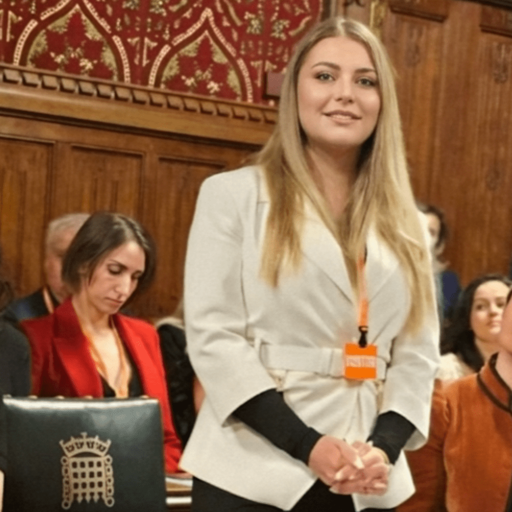 Roxy wears a white blazer and is stood in the House of Commons wearing an orange lanyard