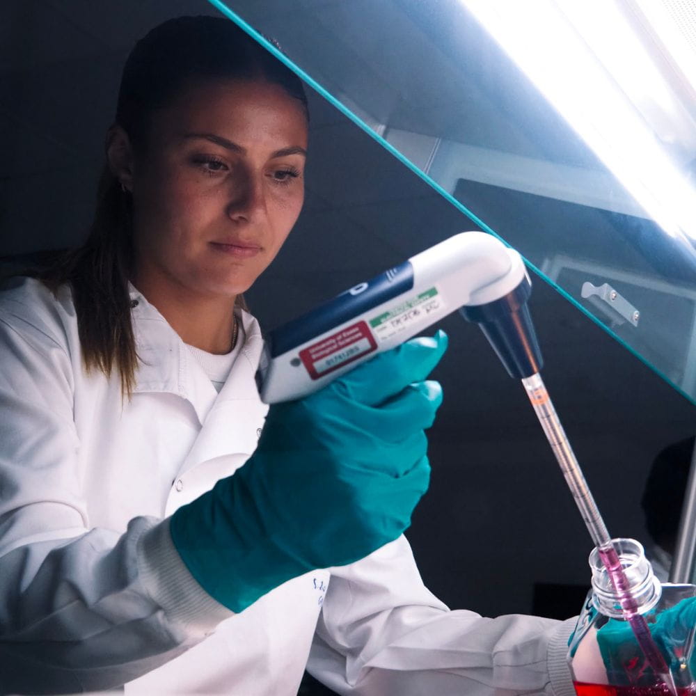 A researcher wearing a white lab coat and blue protective gloves uses an electronic pipette to transfer pink liquid into a sample container inside a laboratory fume hood. Bright overhead lighting illuminates the workspace as the researcher concentrates on the precision task.