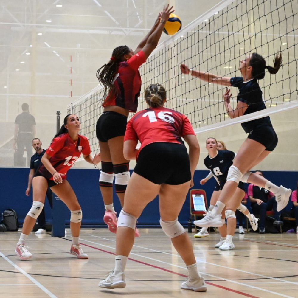 Women’s volleyball players compete at the net, with an England player in red jumping to block an opponent’s attack as teammates prepare for the next play.