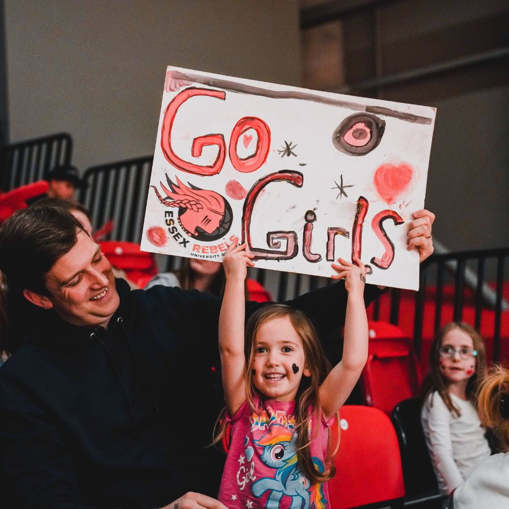 A young girl cheers on the Rebels with her dad, she is holding a sign saying Go Girls