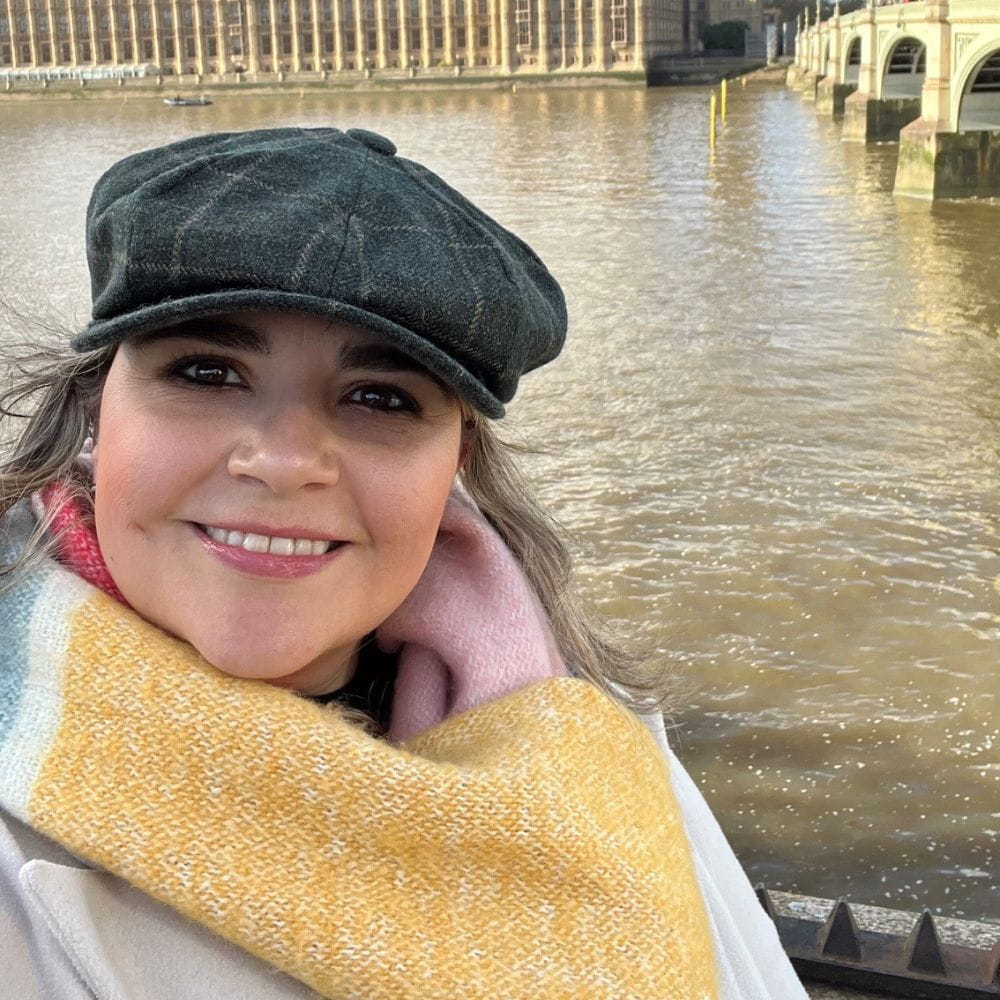 Alba is wearing a scarf and a hat and is stood in front of a river. A bridge and a building can be seen in the background. 