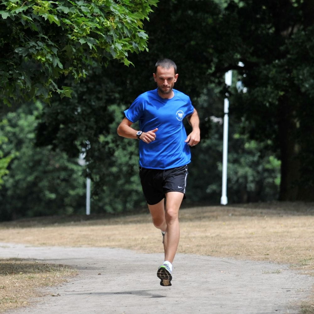 A man in a bright blue T-shirt and black shorts running along a paved path through a leafy park, surrounded by trees and dry grass.