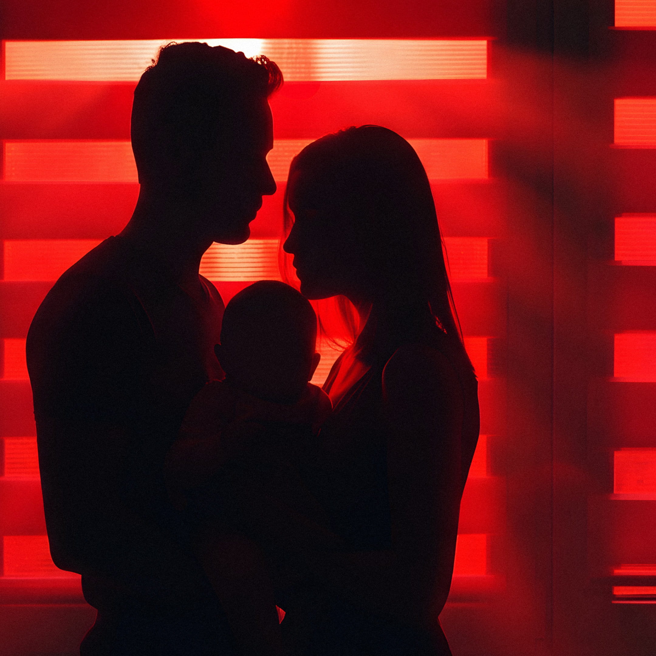 Lightstreams Play promotional image. Silhouette of a man and woman holding a baby. The background is red lights coming through a slatted window