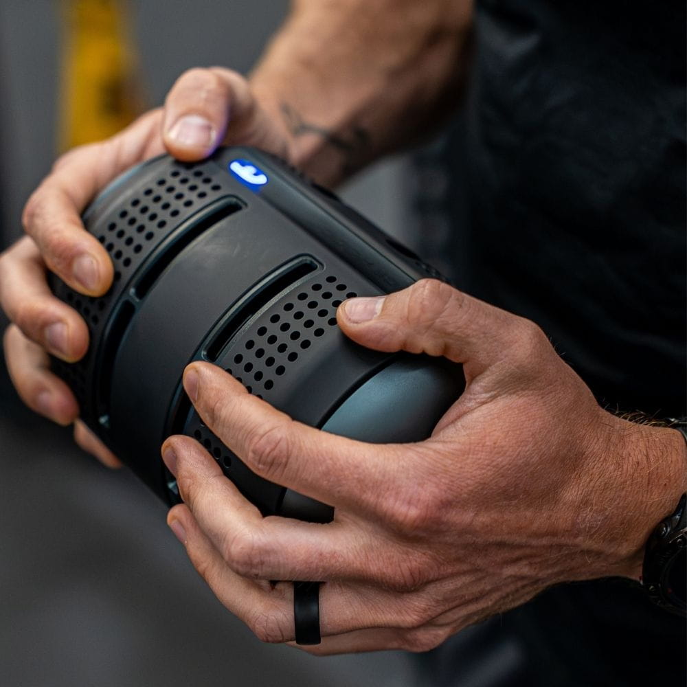 Close-up of a person holding a black palm-cooling device with both hands. The curved device has perforated ventilation holes, recessed hand grips and a blue illuminated power button.
