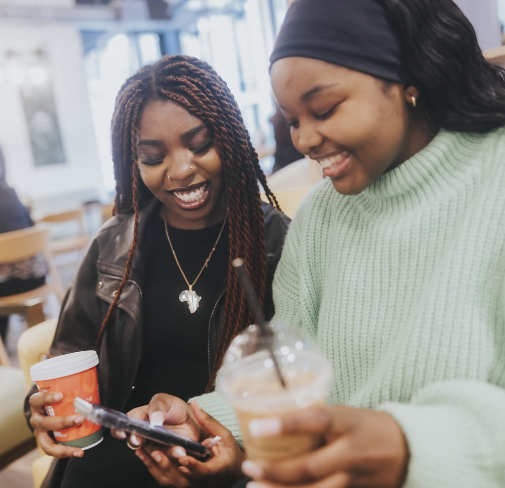 Two young people standing side by side. One is holding a phone which they are both looking at, both are holding drinks cups.