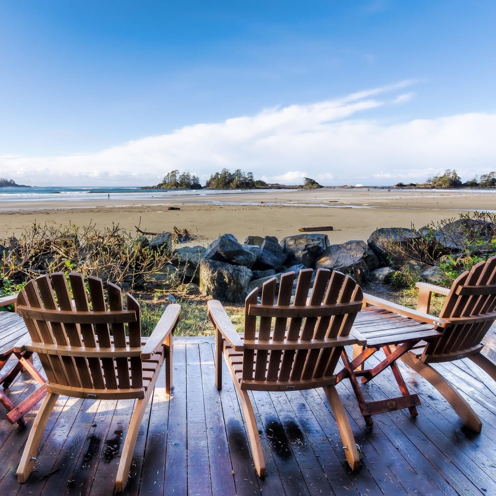 Three wooden chairs on some decking, pointed towards a beach with trees in the background.