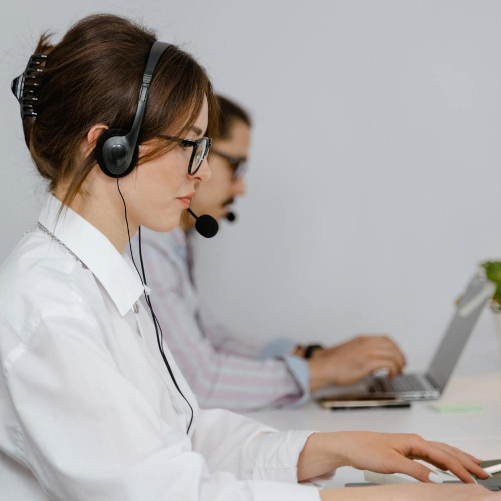 A side-on photo of a woman and a man, both wearing headphones with a microphone, looking at laptops in front of them.