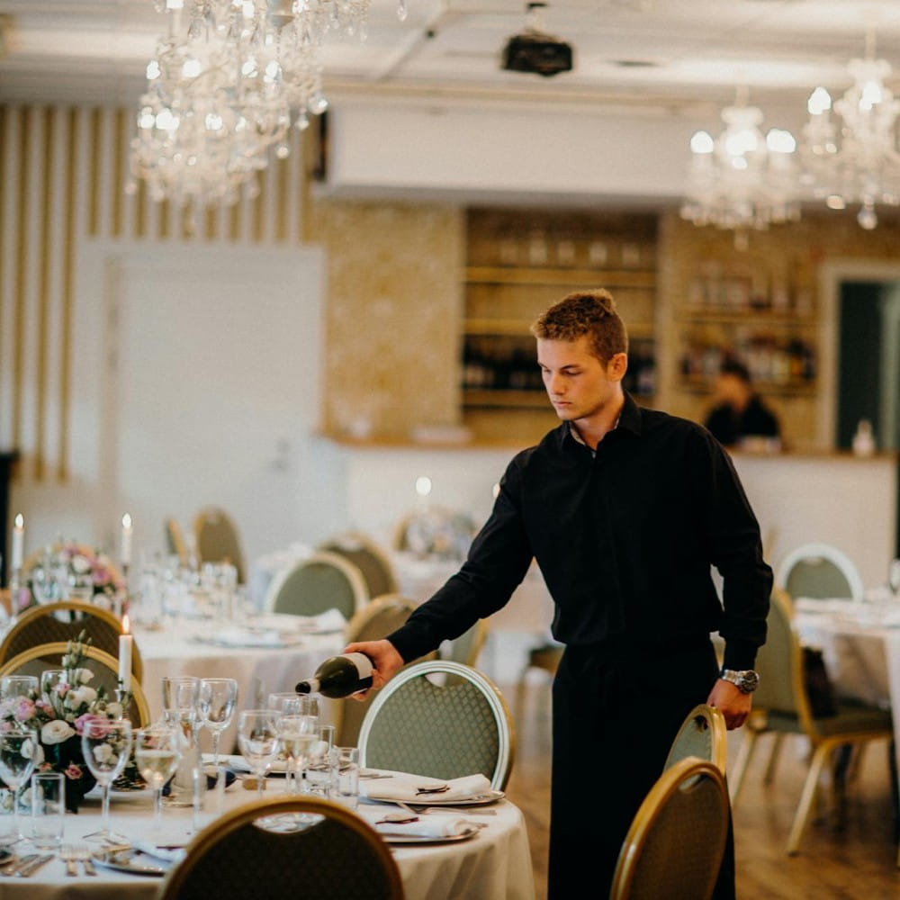 A waiter in smart black clothes pouring wine in a glass. The room is set up for a meal with plates, glasses and flowers on the tables.