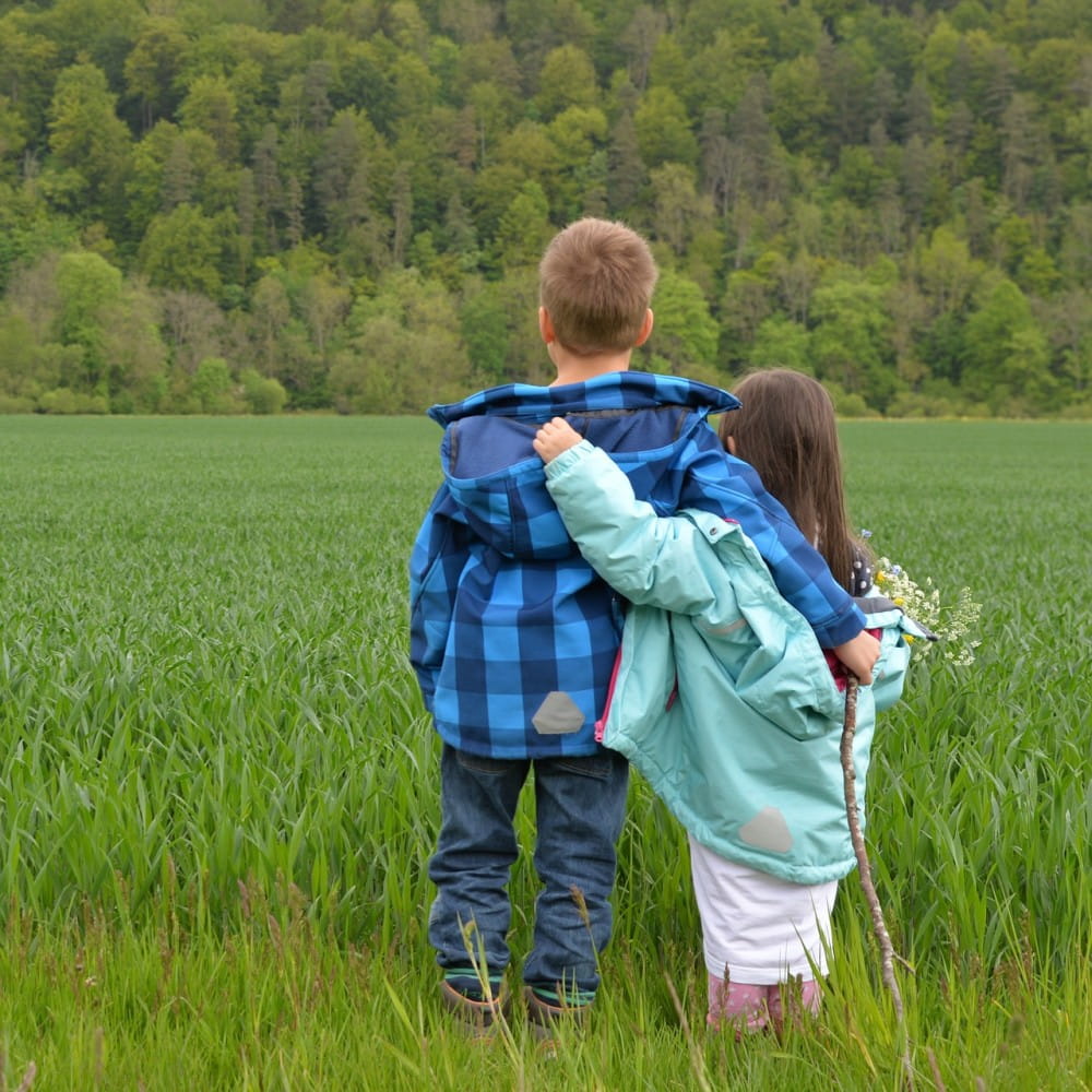 A boy and girl, facing away from the camera, with their arms around each others shoulders as they look across a grass field.