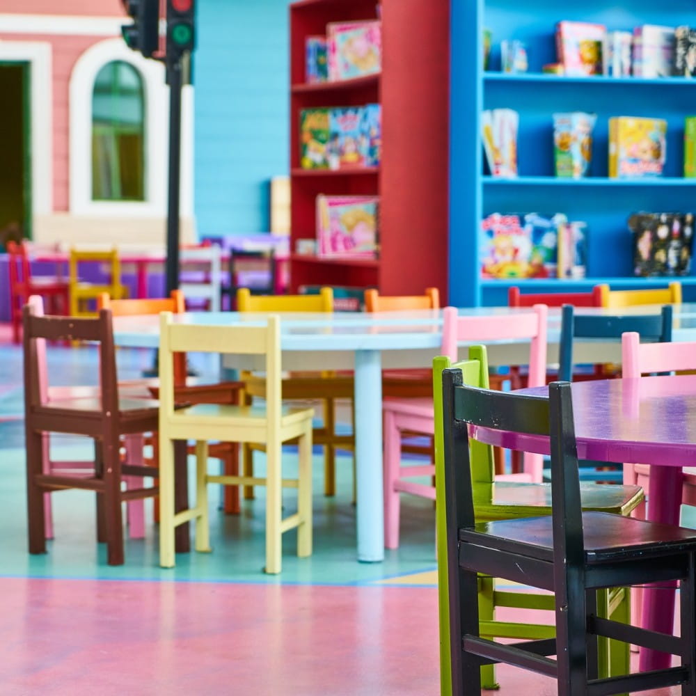 An empty classroom, with small multicoloured chairs placed around tables, with bookcases in the background.