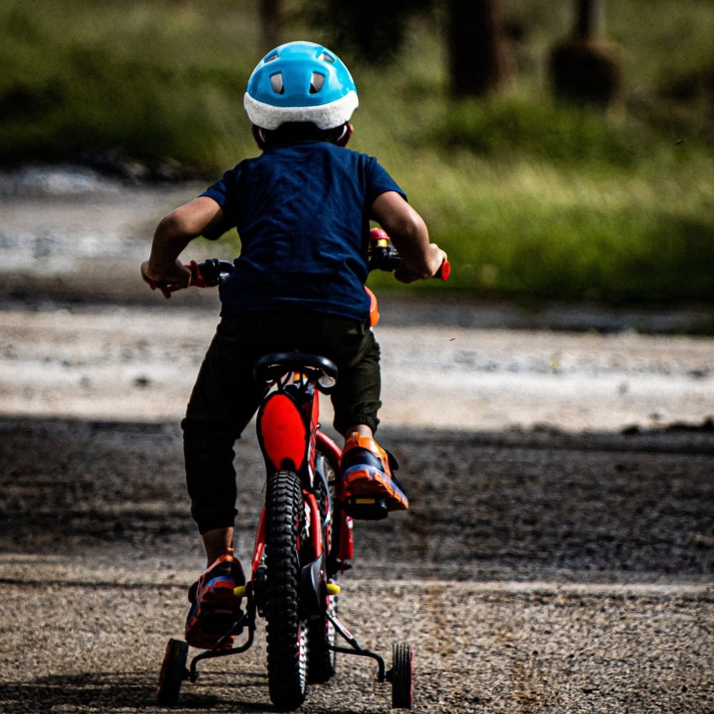 A child wearing a cycle helmet, on a bike with stabilisers, cycling away from the camera.