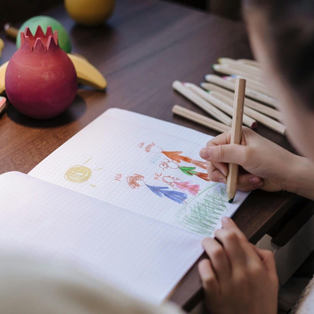 A child, mostly out of shot, using colouring pencils to draw a picture of a family in a notebook.