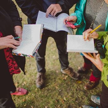 4 students standing outside on the grass with 2 reading notes from books while a third writes