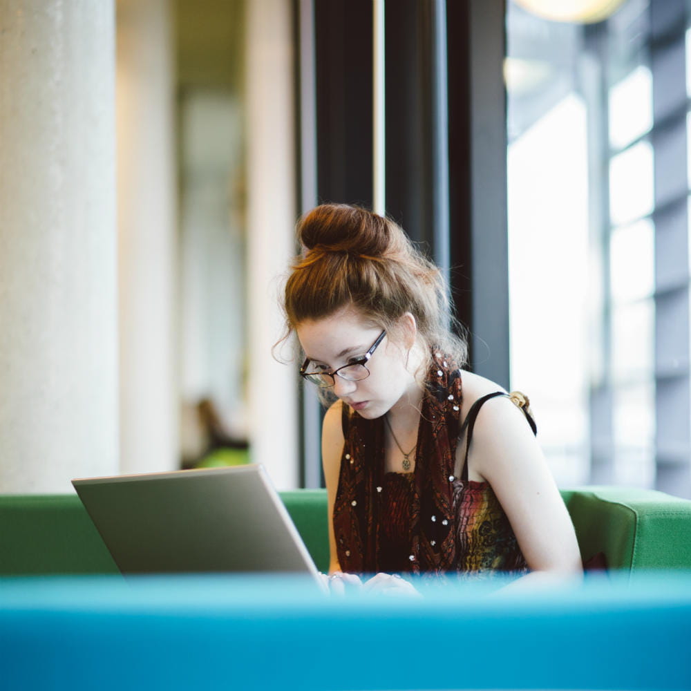 A student sitting on a green chair working on a laptop.