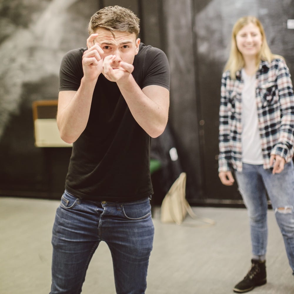 A man pretending to hold a camera and take a photo. A woman is standing in the background laughing.