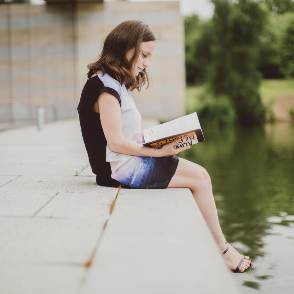 A woman reading a book, sitting on a step next to a lake with her feet hanging over the edge.