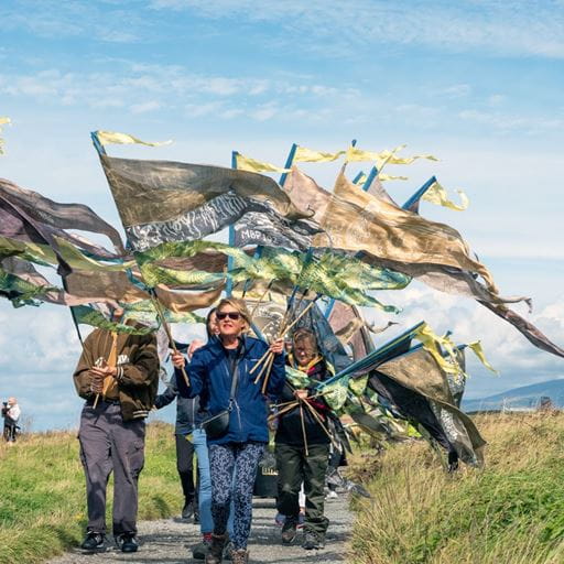 A group of people carrying large, colourful flags which are flapping in the wind, walk along a coastal path with a clear sky behind