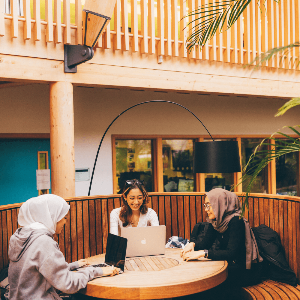 Three female students sat in a round wooden seating area with a balcony in the background