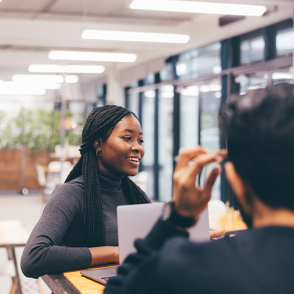 A black female student sits at a table in the University of Essex's Innovation Centre cafe with her Apple laptop. She has a warm and infectious smile and her long braids hang over her shoulder and down her grey turtleneck sweater. She is in conversation with a male student who is facing towards the female student, we cannot see his face but he had black hair, a black beard and a black sweater. 