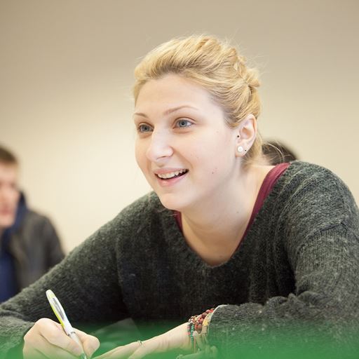 A smiling female student holding a pen and writing in a notepad whilst sat at a desk.