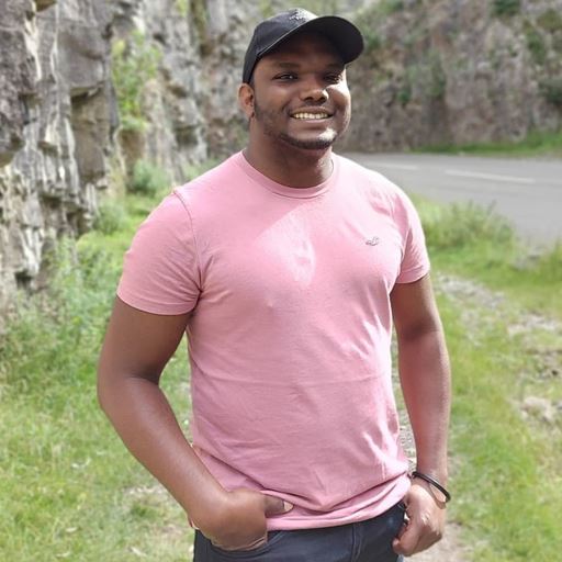 Life Sciences graduate Akira Williamson, standing outside wearing a pink t-shirt and dark blue baseball cap. He is smiling at the camera, with some cliffs and a road visible in the background.