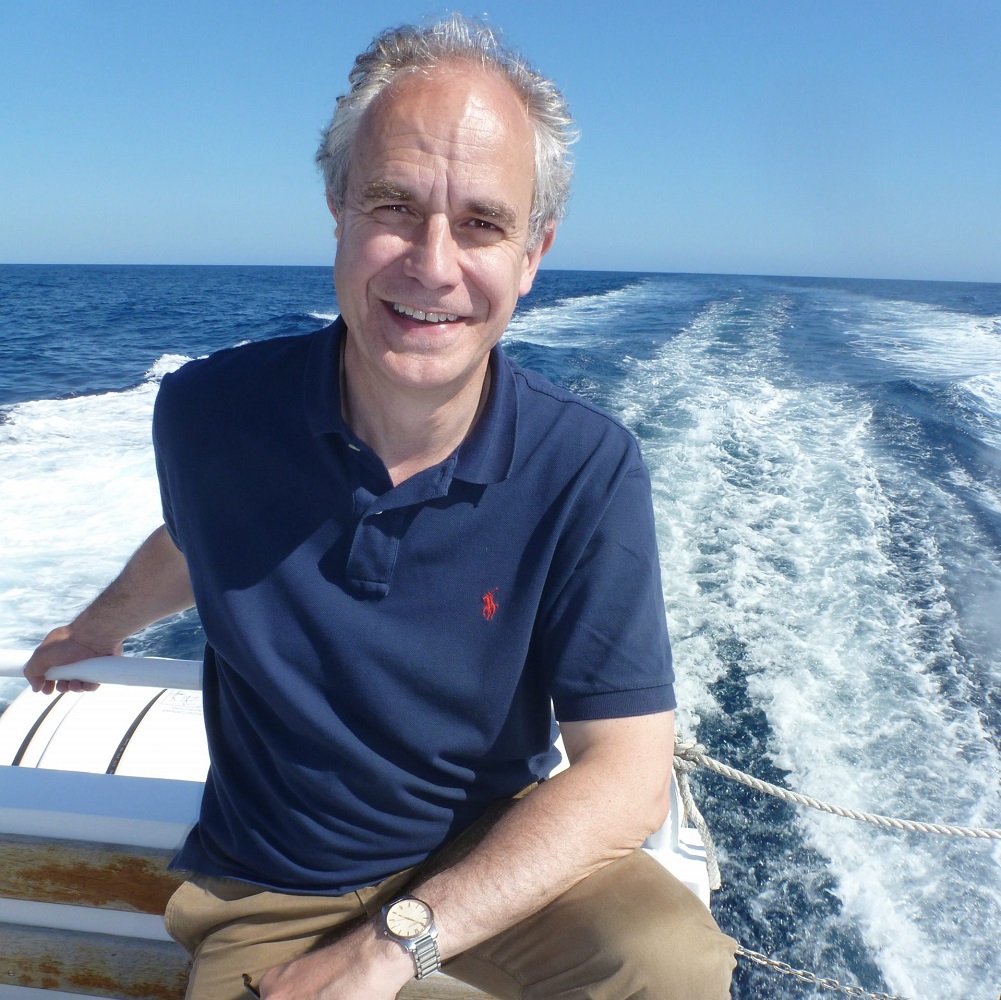 Charles Clover, sitting on the back of a boat in a blue t-shirt, with the sea and blue sky in the background.