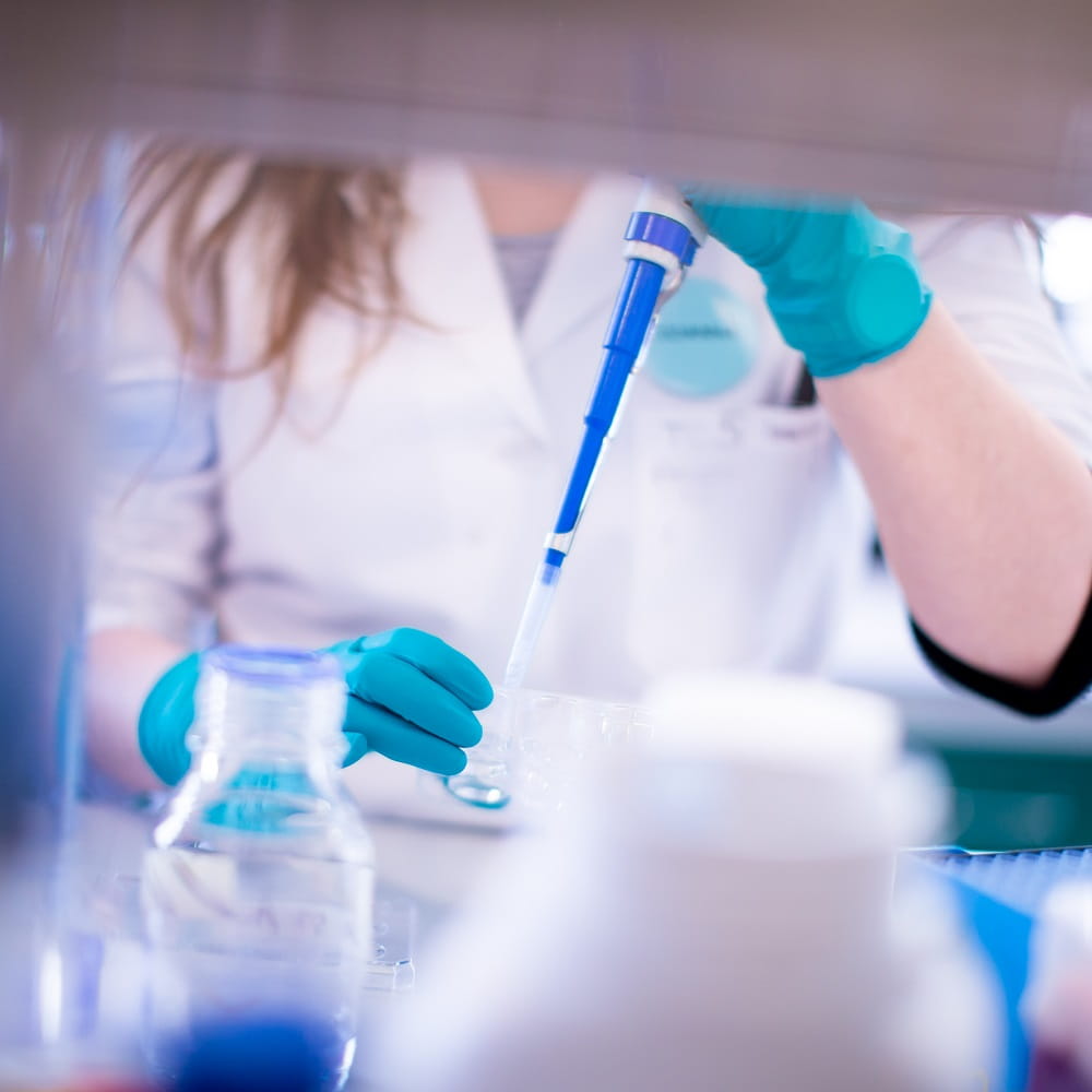 Taken through a gap between some shelves. You can see the torso and arms of a person with long hair, wearing teal-coloured gloves, using a pipette to put something into a beaker.