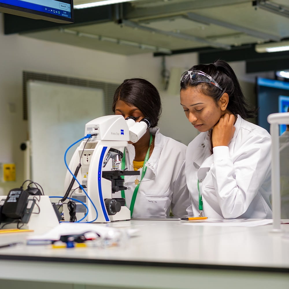 Two female students wearing white coats, sitting at a lab bench with a microscope in front of them. They are looking down at an iPad on the bench top.