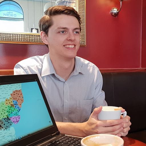 A photo of CSEE graduate Andrew Starkey, holding a cup of coffee. There is a red wall and the corner of a large rectangular mirror behind him.