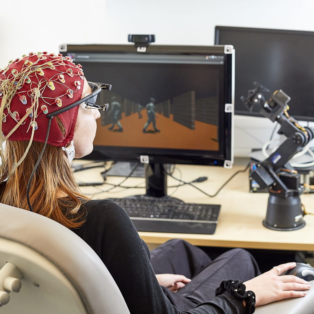 A woman with long hair wearing an EEG cap, sitting in a chair in front of a desk with a computer monitor and a small robotic arm in front of her.