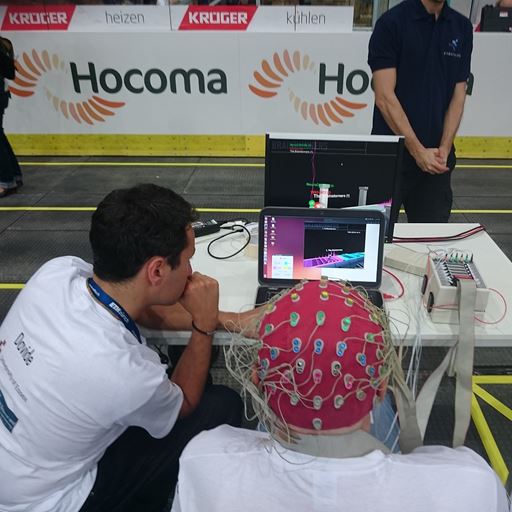 A person sitting in front of a laptop wearing an EEG cap, with someone crouched to their left working on the laptop.