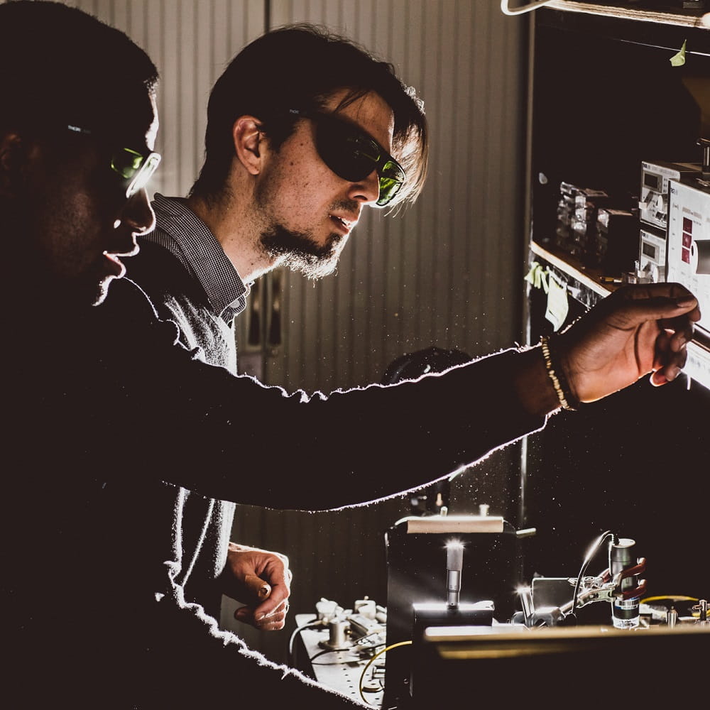 Two men wearing tinted eye protector glasses, one of them reaching out to a switch on a piece of equipment on a shelf at head height.
