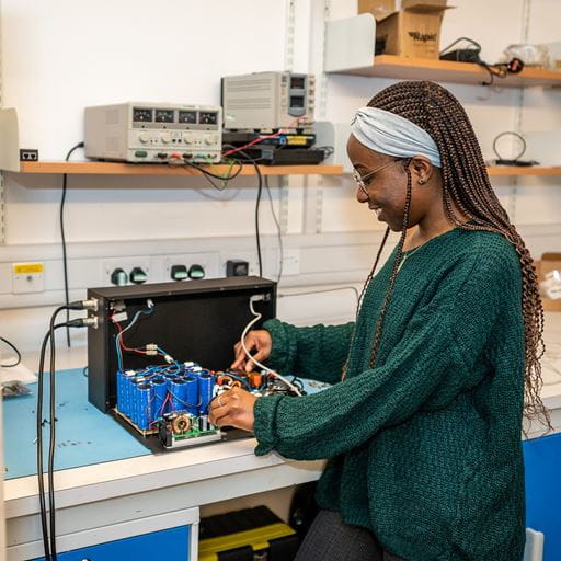 A young Black woman in a green top is working on some wires in an acoustic device that is sitting on a lab bench.
