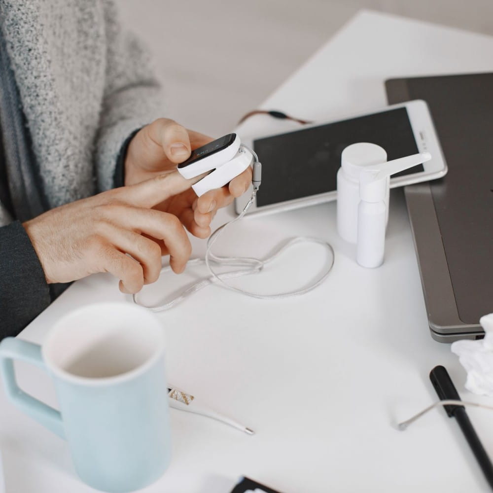 A person with a digital monitor on their finger. A phone, a mug and tissues are on the table in front of them.