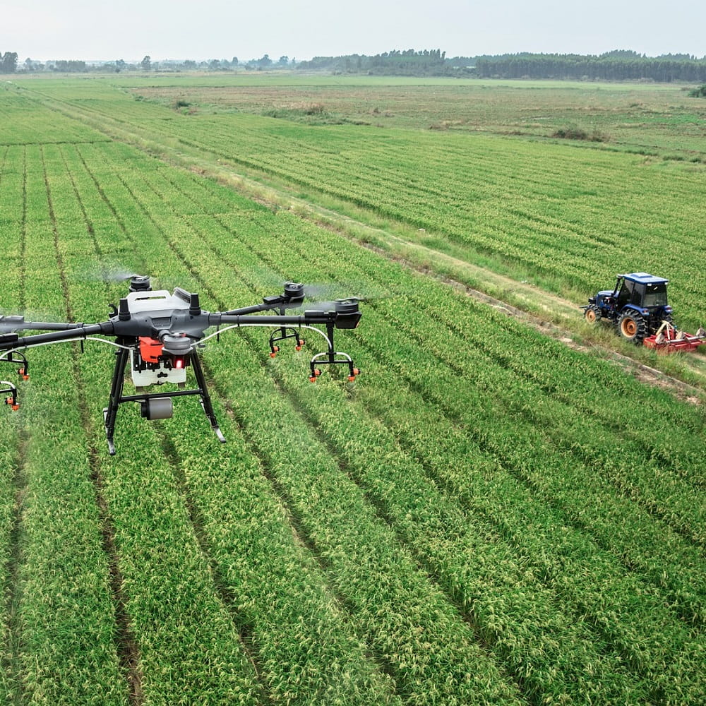 An aerial shot of a field. On the left is a drone hovering in the air, on the right a tractor is ploughing the field.