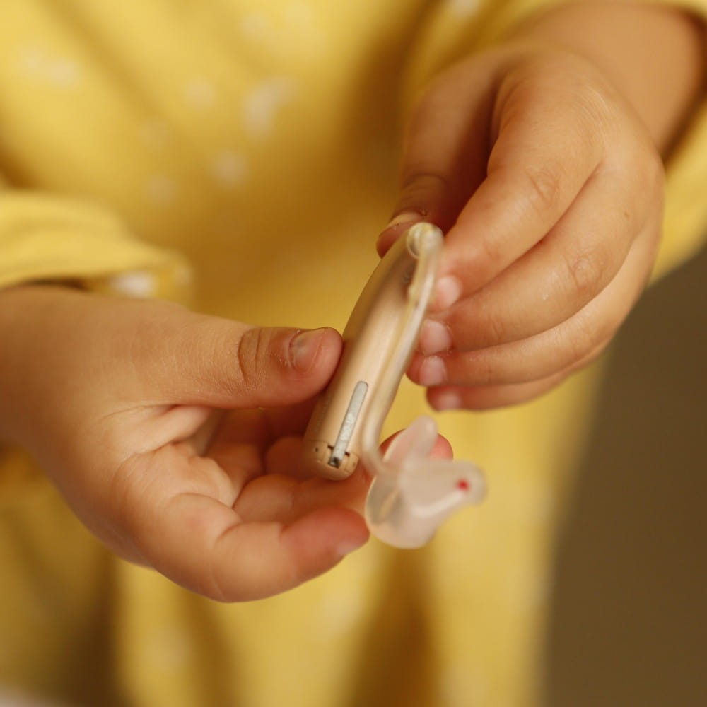 A small child in a yellow top, mostly out of shot, holding out a hearing aid in both hands.