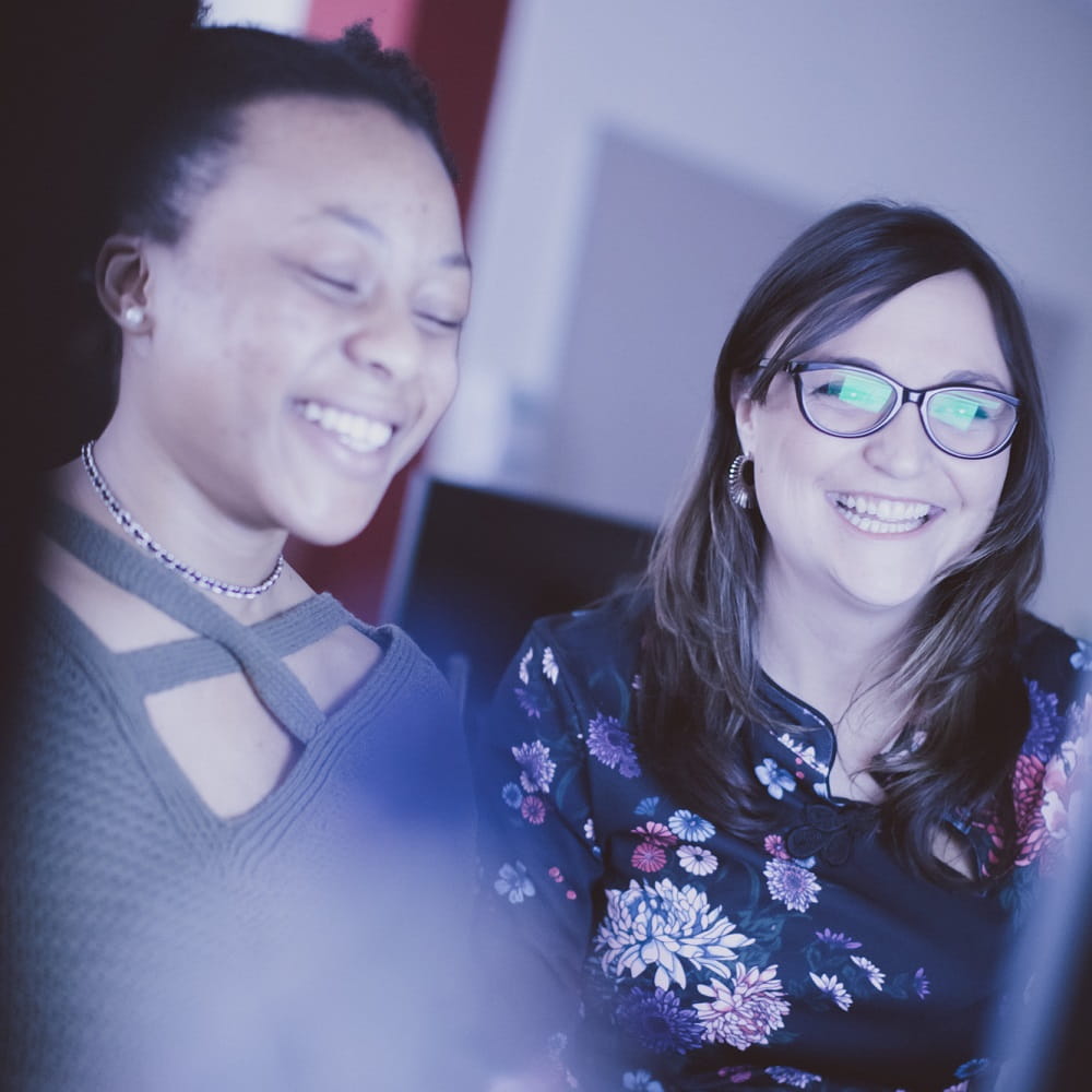 Dr Alba Garcia and a female student working at a screen together, the student is smiling and has her eyes closed. Alba is wearing glasses which are reflecting some of the screen. The photo has a pale blue tint to it.