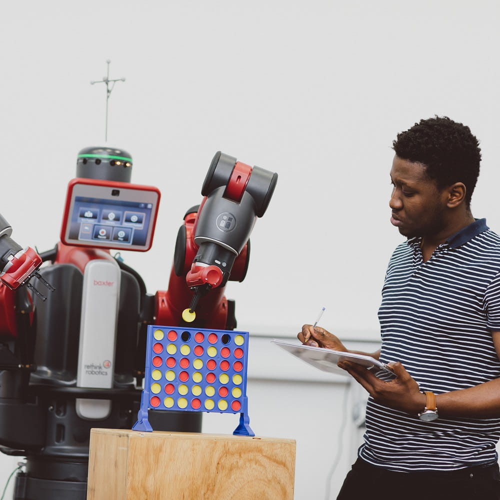A young black man holding an iPad and stylus, standing to the right of a large red Baxter robot that is playing Connect-4.