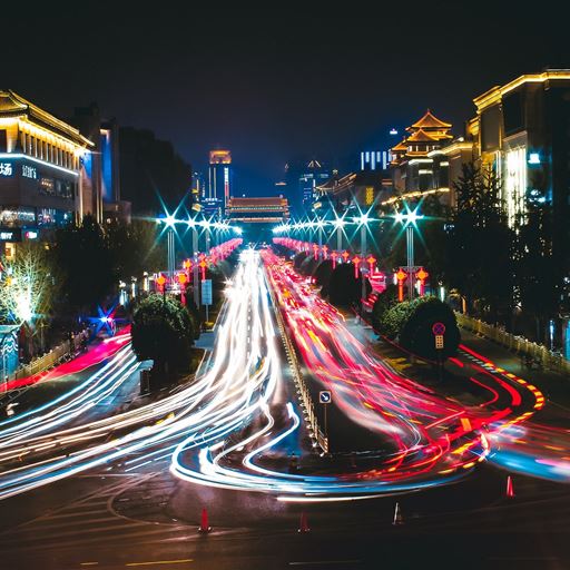 A night shot of a road in Xi'an City, China. Car headlights and brakelights are in the middle of the shot as people drive up and down a road, while on both sides buildings are lit up.