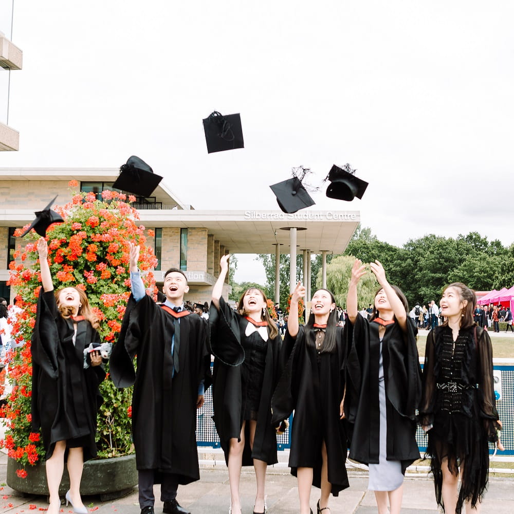 A row of six graduates standing outside, wearing graduation gowns and looking up as they throw their mortarboards up in the air.
