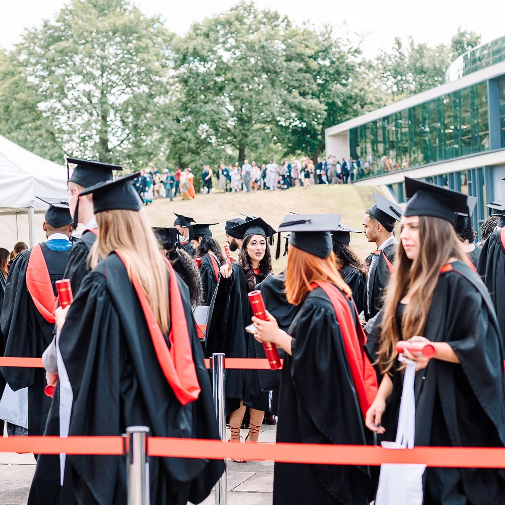 In the middle is a graduate standing in a queue, talking to three other graduates in the foreground with their backs to the photographer. A line of people outside a building can be seen in the background.